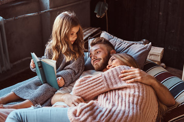 Mom, dad and daughter reading storybook together while lying on bed.