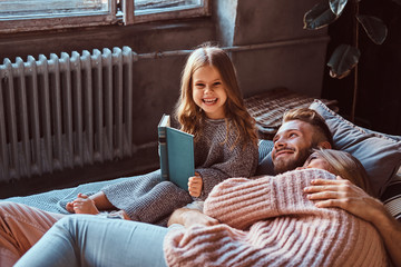 Wall Mural - Mom, dad and daughter reading storybook together while lying on bed.