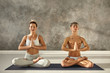 Indoor portrait of two barefoot young people man and woman with flexible strong bodies meditaing on one mat during yoga class, sitting in lotus posture, closing eyes and holding hands in namaste