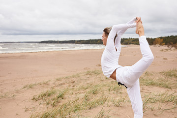 Wall Mural - Cropped side view of sporty young man working out outdoors, training balanca and flexibility, doing backbend, standing in Natarjasana on sand. Male yogi maintaining balance, doing Dancer King pose