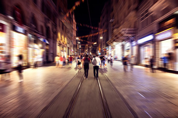 Blurry motion image of people walking in Istiklal Avenue (the city’s main pedestrian boulevard) at night in Istanbul. The street which is lined with 19th-century buildings, shopping chains and cafes.