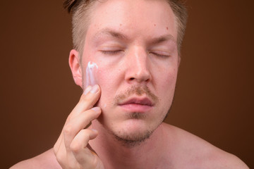 Young handsome man shirtless while grooming against brown backgr