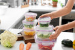 © Pixel-Shot - Woman holding stack of plastic containers with fresh vegetables for freezing at table in kitchen