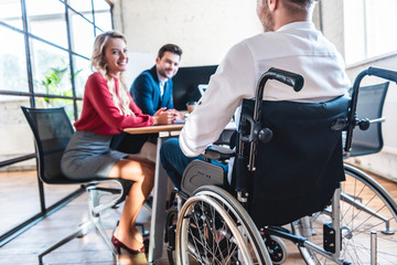 Wall Mural - partial view of smiling businesspeople looking at colleague in wheelchair in office