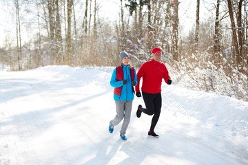 Wall Mural - Mature spouses in winter sportswear jogging down snow road on sunny day in the forest