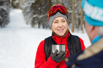 Wall Mural - Happy mature woman in activewear holding cup of hot tea while talking to husband after skiing training