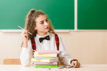 schoolgirl in school uniform sitting at her desk with books and pencils on the background of a green board