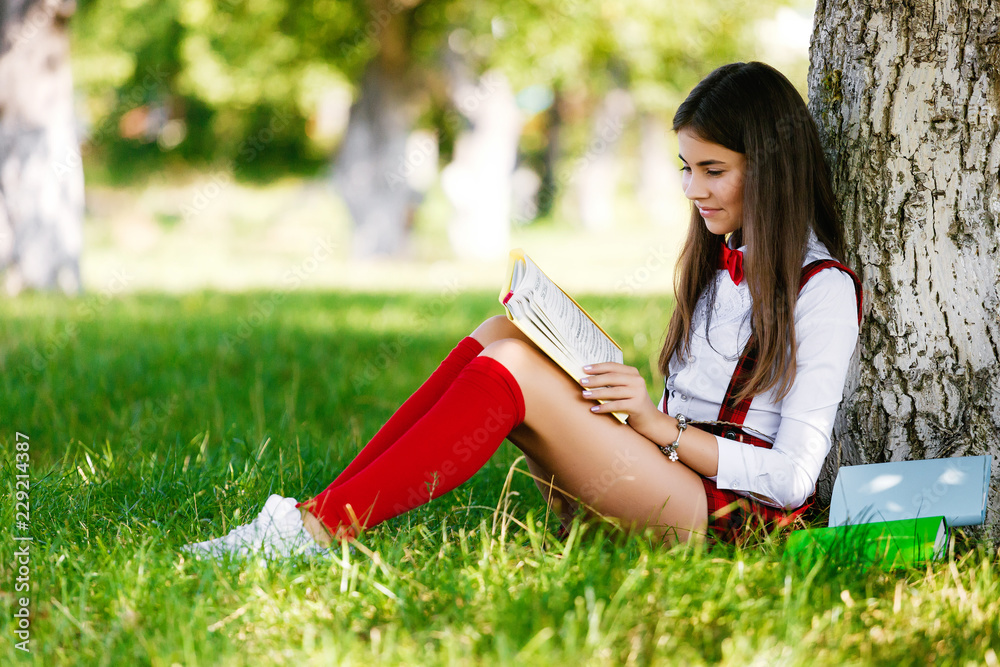 young schoolgirl in school uniform sitting on the grass under a tree ...
