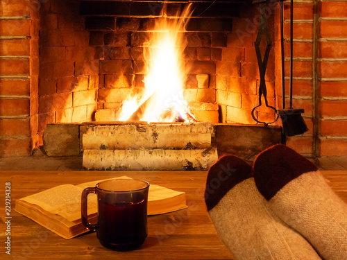 Cuadros en Lienzo Cup of tea, book, women's feet in warm socks on a wooden table opposite a burnin