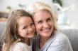 Portrait of happy grandmother and granddaughter spend time at home together, hugging and cuddling on couch, smiling granny embrace cute little grandchild looking at camera making family picture