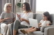 © fizkes - Small girl sit alone using laptop while mom and granny talk aside, having adult conversation, little daughter entertain herself watching video on computer, mother and grandmother chatting