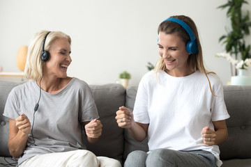 Funny elderly mother and adult daughter listen to music in earphones having fun at home together, happy young woman enjoy spending time with aged mom, hear favorite tracks in headphones