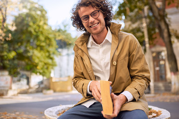 Poster - Happy handsome young man reading and posing with book outdoors. College male student carrying books in campus in autumn street. Smiling smart guy wears spectacles and curly hair reading books outside