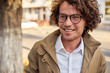 Closeup portrait of young businessman with glasses smiling and posing outdoors. College male student in autumn street. Smart guy in casual wears spectacles with curly hair walking down the street