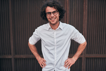 Outdoor shot of smilimg handsome young businessman with glasses posing outdoors. Male student posing against brown wall. Smart guy in white shirt wears spectacles with curly hair walking on street