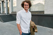 Handsome young happy businessman with glasses smiling and posing outdoors. Male student in autumn street. Smart guy in white shirt wears spectacles with curly hair walking on street