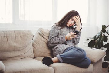 Frustrated woman reading message in a smartphone. Upset girl sitting in living room holding mobile phone. Bad news, emotions, feelings, conversation concept