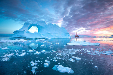 little red sailboat cruising among floating icebergs in disko bay glacier during midnight sun season