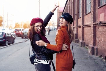 Outdoors fashion portrait young pretty best girls friends in friendly hug. Walking at the city. Posing at the street. Wearing stylish outerwear and hats. Bright make up. Positive emotions.