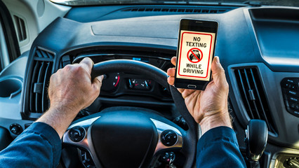 Close up of the hands of a man driver, holding phone in hand while driving, phone displays no texting while driving on screen. Very unsafe driving habit.