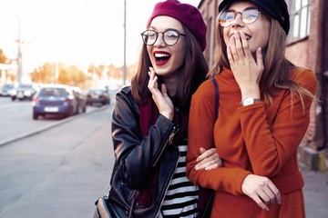 Outdoors fashion portrait young pretty best girls friends in friendly hug. Walking at the city. Posing at the street. Wearing stylish outerwear and hats. Bright make up. Positive emotions.