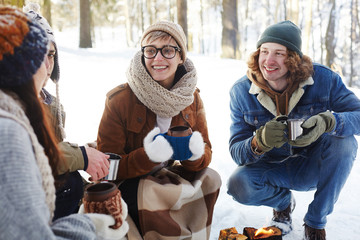 Portrait of happy young people camping in winter forest sitting in circle round fire and chatting while holding cups