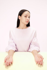 Wall Mural - Serious business woman sitting at table on a pink studio background. The portrait in minimalism style