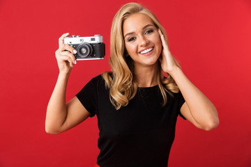 Poster - Portrait of young blond woman 20s smiling and photographing on retro camera, isolated over red background in studio