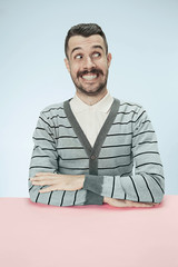 Wall Mural - The happy and smiling business man sitting at table on blue studio background. The portrait in minimalism style