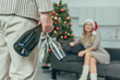 © LIGHTFIELD STUDIOS - cropped shot of man with champagne glasses standing in front of woman in santa hat sitting on couch at home on christmas