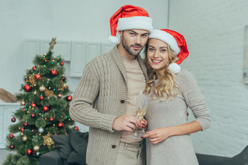 Wall Mural - happy young couple with champagne glasses looking at camera in front of christmas tree at home