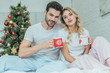 © LIGHTFIELD STUDIOS - beautiful young couple with cups of cocoa sitting in bed at home on christmas and looking at camera