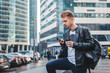 © olezzo - Attractive stylish young man in a leather jacket with a smartphone and take-out coffee in his hands against the backdrop of a large modern city