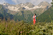 © Лариса Левина - Young woman with black hair in a long red dress on the background of snow-capped mountains in a green valley in the summer.