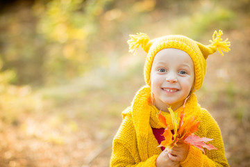 Wall Mural - Cute little girls with big bue eyes playing on beautiful autumn day. Happy children having fun in autumn park. The girl is dressed in yellow coat and a hat. She holds a bouquet of yellow leaves in her