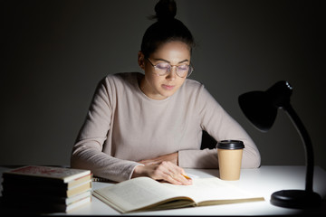 Busy female teenager reads book, uses table lamp, prepares for final examination, sits at desktop, wears optical round glasses, poses against dark background. Reading in evening. Studying concept