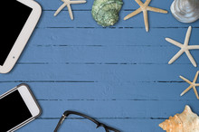 BACKGROUND OF SEASHELLS, TABLET, SMARTPHONE, AND A PAIR OF GLASSES ON BLUE OIL PAINTED WOODEN TABLE. COPY SPACE
