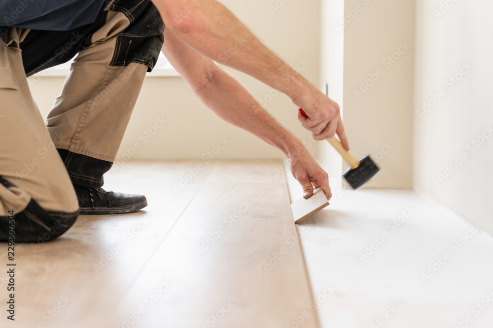 Strikes with a soft hammer on the part with a lock, for fixing. Installing laminate flooring fitting the next piece - focus on hand. Man laying laminate flooring - obrazy, fototapety, plakaty 