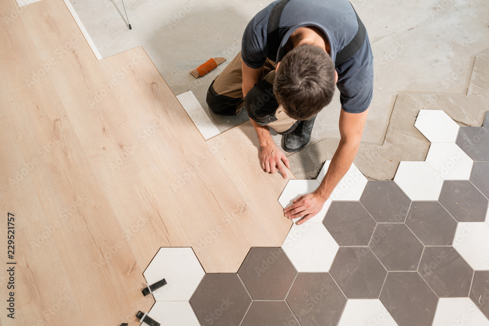 Male worker installing new wooden laminate flooring. The combination of wood panels of laminate and ceramic tiles in the form of honeycomb. Kitchen renovation. - obrazy, fototapety, plakaty 