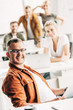 © LIGHTFIELD STUDIOS - smiling young man working at open space office and looking at camera with colleagues on background