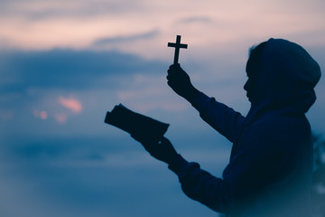 Wall Mural - Silhouette of teen girl hands holding wooden cross and bible on sunrise background, Crucifix, Symbol of Faith.