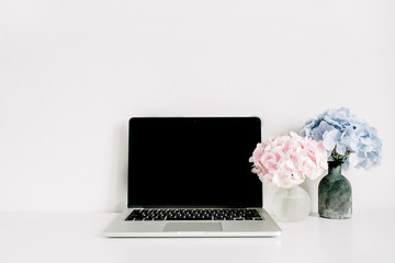 Blank screen laptop and pink and blue hydrangea flower bouquets on white background. Minimal home office desk workspace.