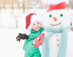 Wall Mural - portrait of smiling boy in red christmas hat holds gift box and hugs snowman