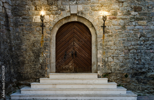 Fotomural Old wooden doors of ancient castle with white staircase are illuminated by burning torches