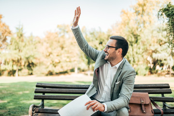 Elegant young man sitting on a bench and waving at a friend.