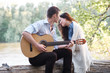 © soleg - young couple sitting on a log by the river and playing guitar, summer nature, bright sunlight, shadows and green leaves, romantic feelings