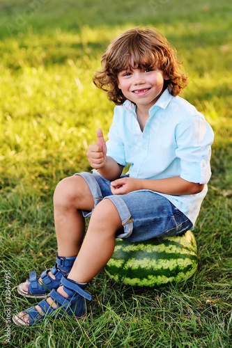 Cute Baby Boy With Curly Hair Sits On A Big Watermelon And Smiles