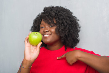 Wall Mural - Young african american woman over grey grunge wall eating green apple with surprise face pointing finger to himself