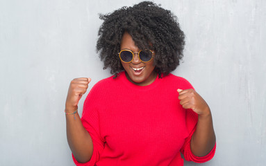 Sticker - Young african american plus size woman over grey grunge wall wearing retro sunglasses very happy and excited doing winner gesture with arms raised, smiling and screaming for success