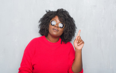 Sticker - Young african american plus size woman over grey grunge wall wearing retro sunglasses pointing finger up with successful idea. Exited and happy. Number one.